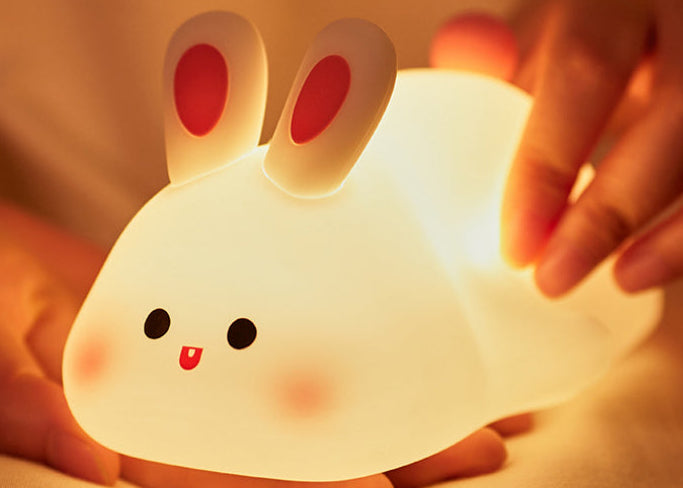 White silicone rabbit night light glowing warm on a bed, with a hand resting beside it