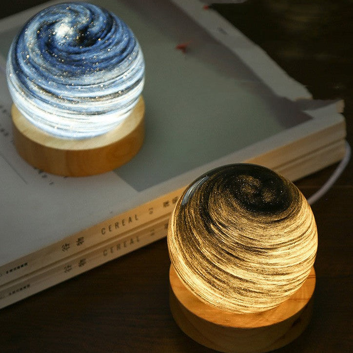 Two planet night lights side by side, one blue and one gold, glowing on their wooden bases beside a book